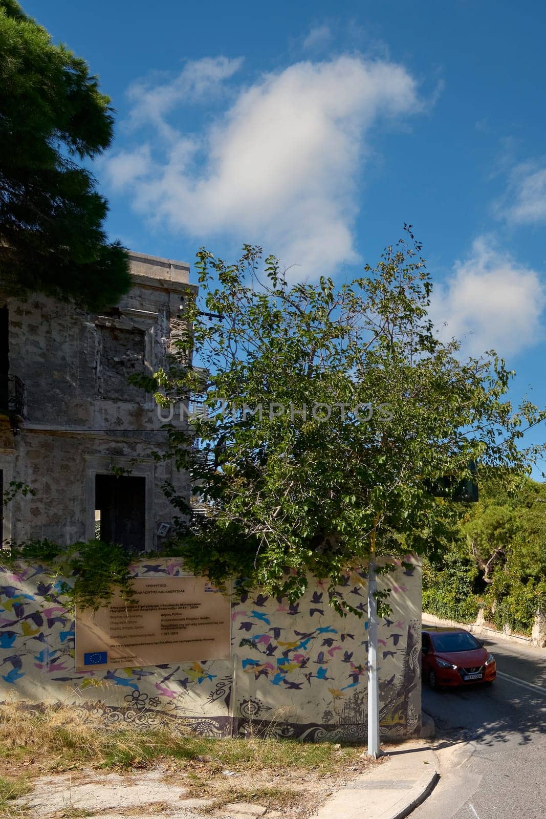 Rhodes. Greece - June 06, 2025: Urban scene capturing a vibrant mural decorating an old wall in Rhodes, with a lush tree in the foreground and a vehicle passing by. The combination of art and historic