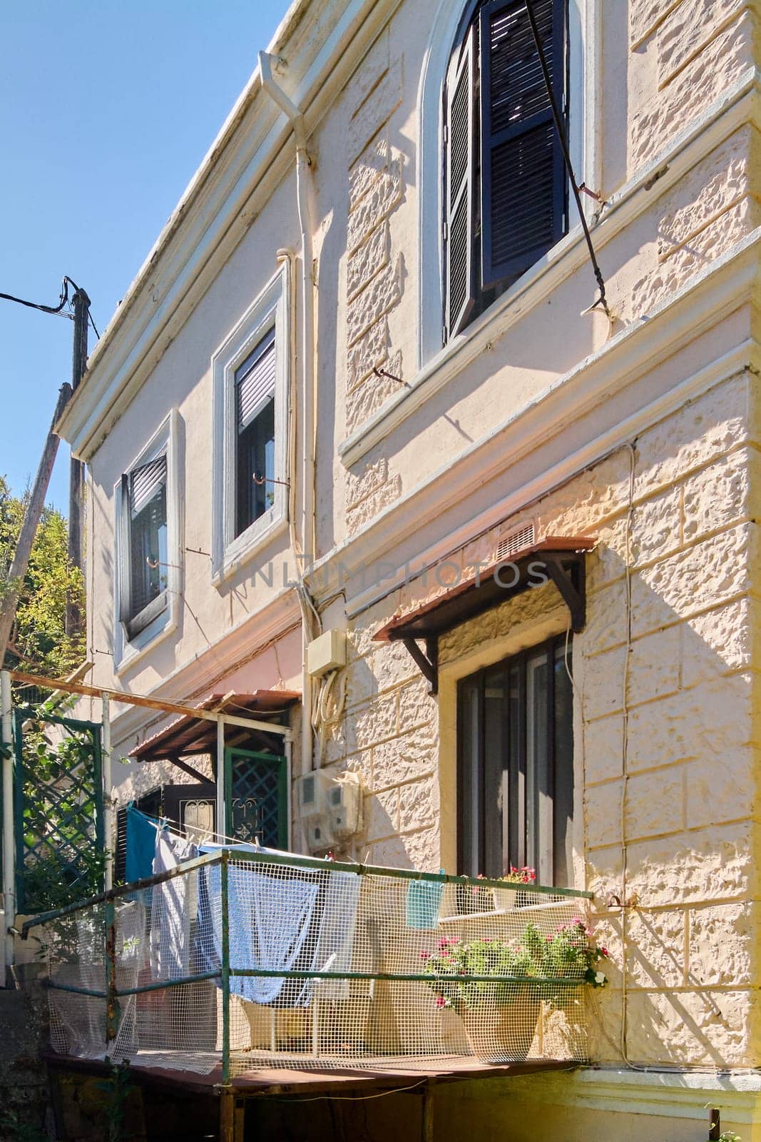 Detail of a traditional house facade in Rhodes, showing a balcony with laundry, flower pots, and dark shutters. It reflects the authenticity of Mediterranean daily life under the sunlight.