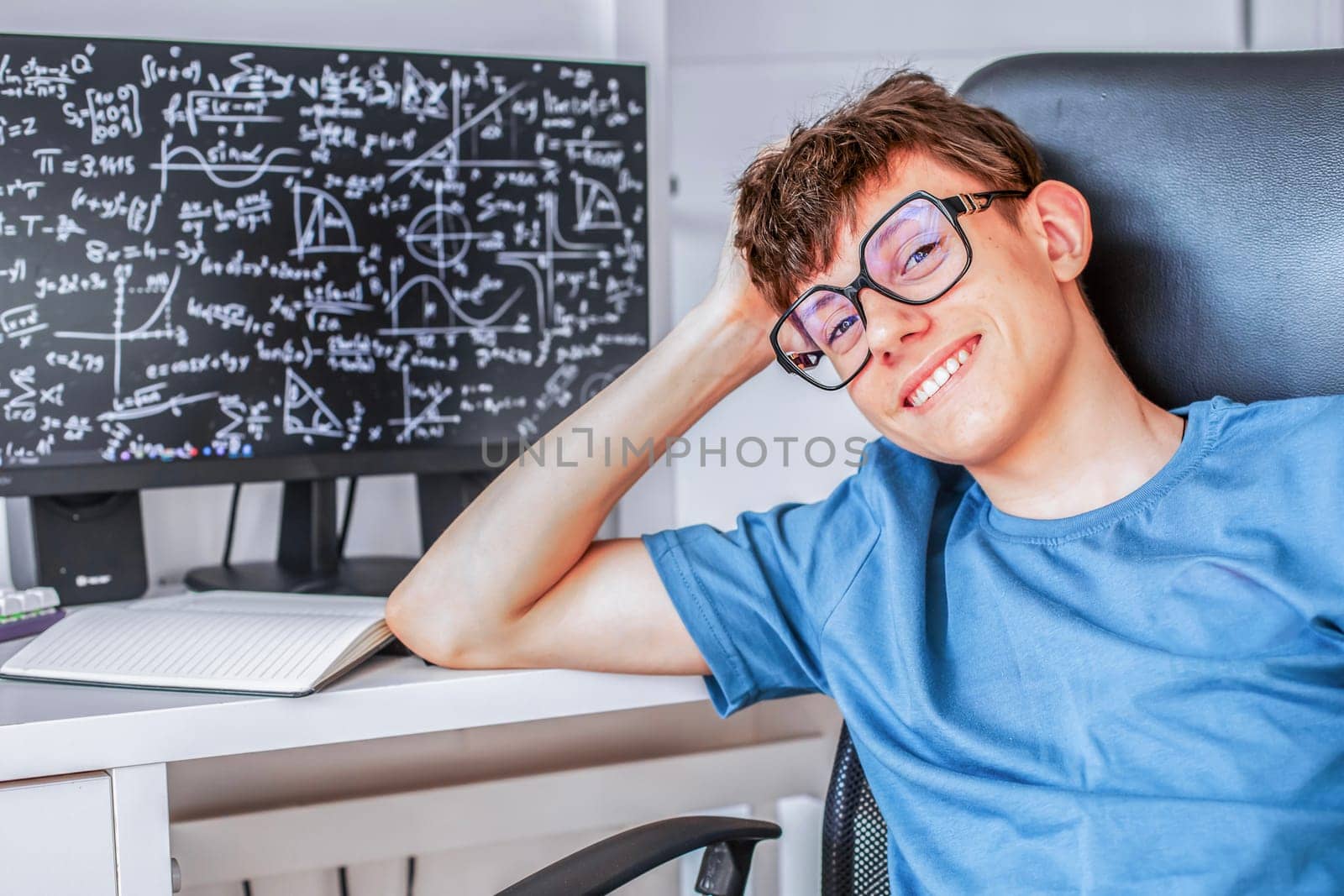 Cute teenage student in glasses sitting at a computer studying online, looking at the camera and smiling contentedly. High quality photo