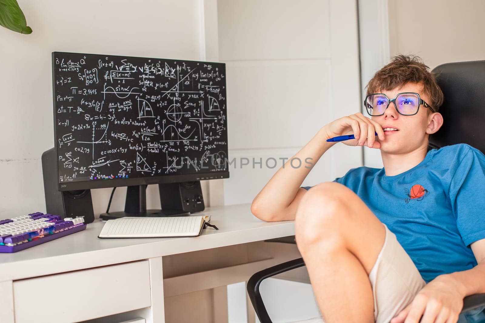 A teenage boy in glasses ponders solving math problems while sitting at a computer monitor. High quality photo