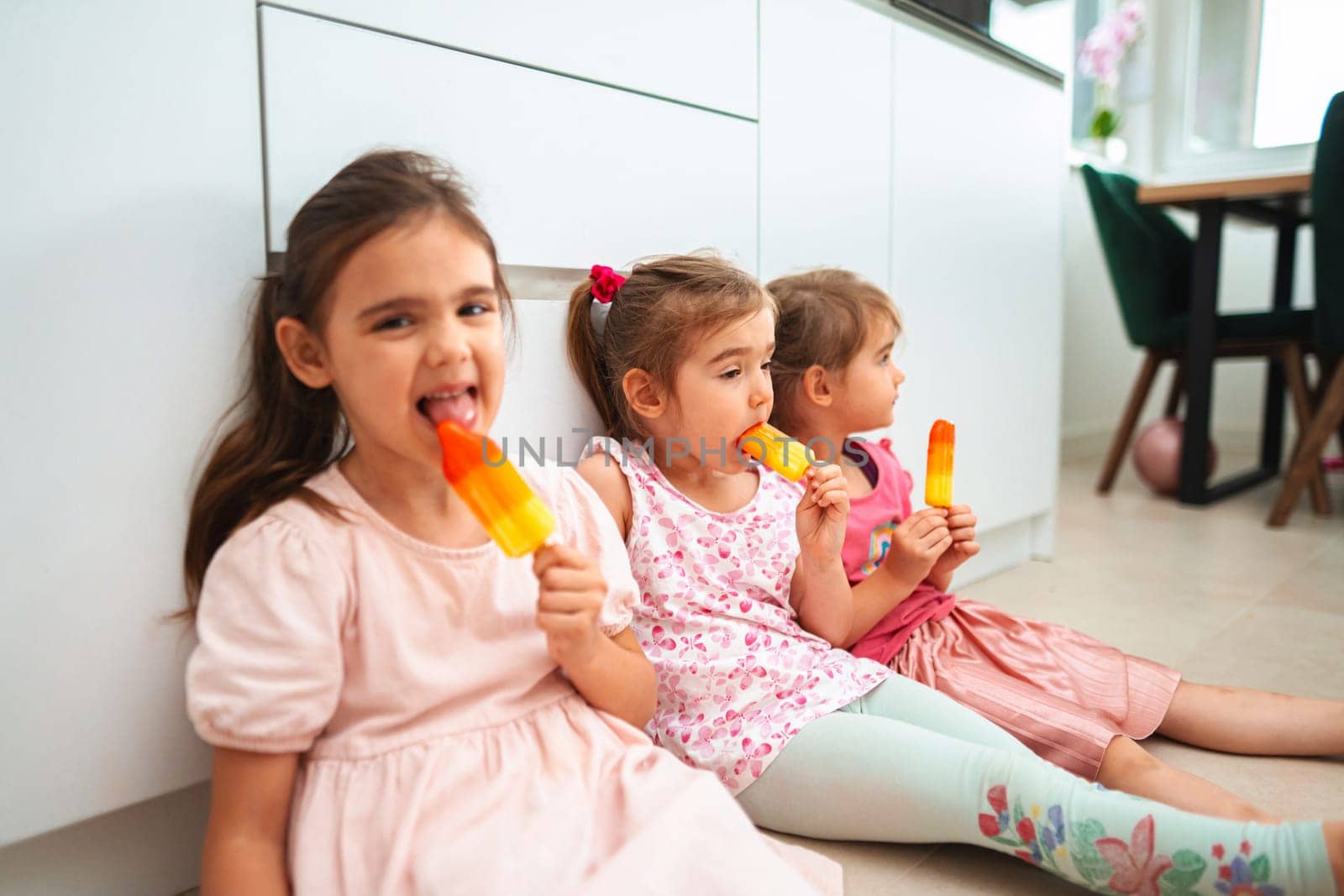 Three little girls sit on a floor, savoring bright ice pops.
