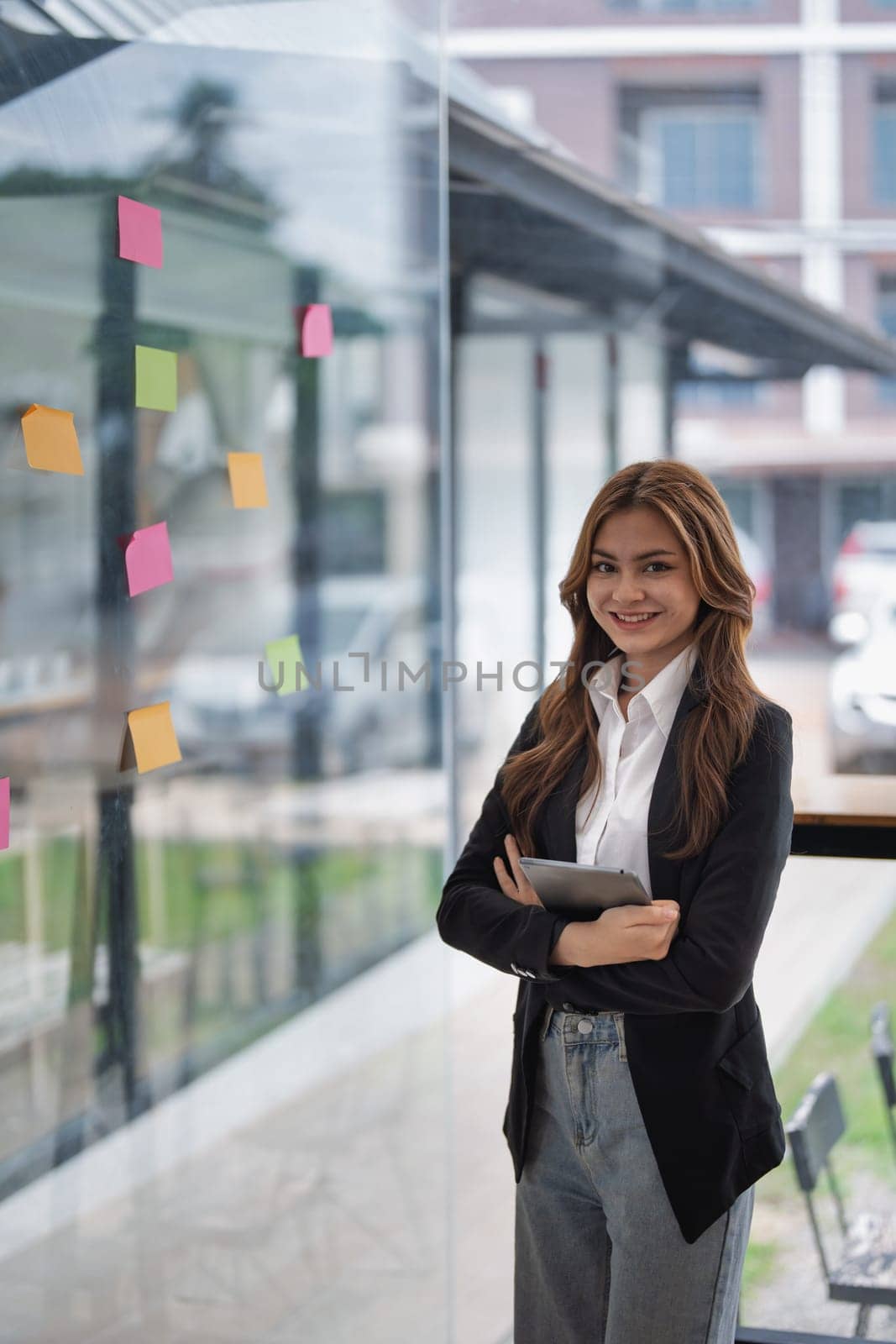 Smiling businesswoman holds tablet while standing by window with notes.