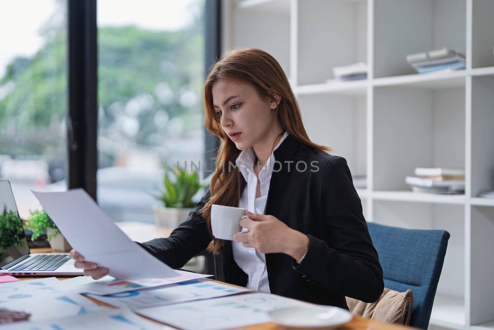 Young businesswoman analyzes paperwork while drinking coffee at her desk.