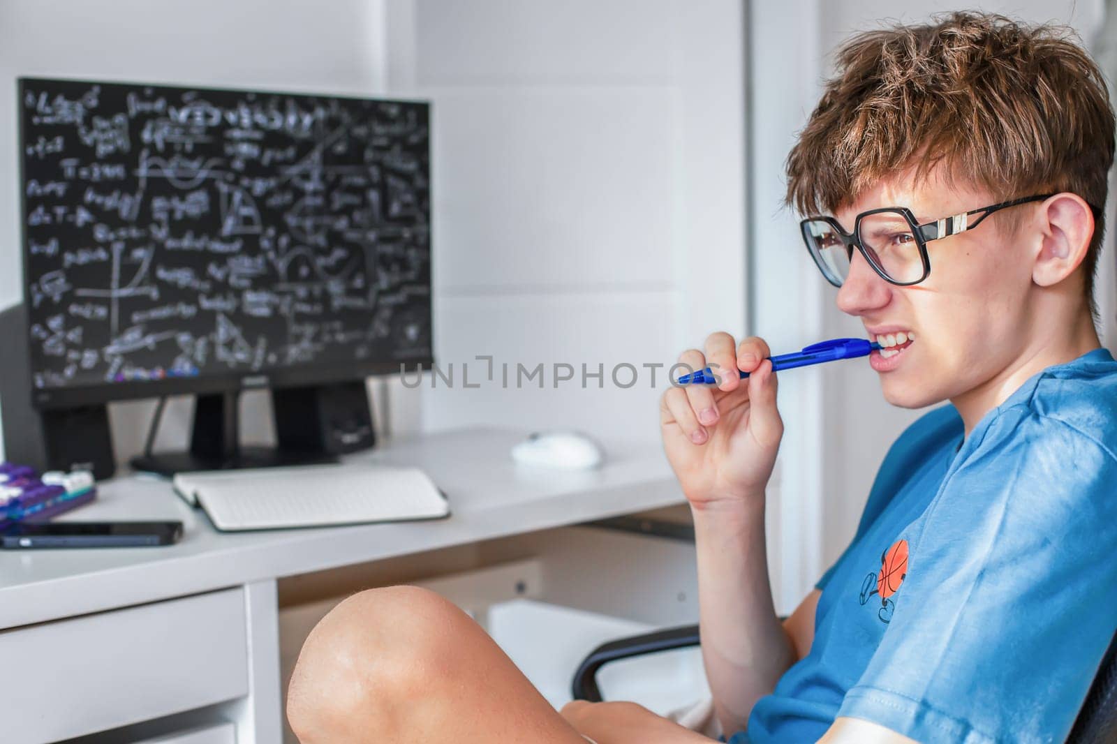 A student in glasses sits studying at a computer monitor with a pen in his teeth, and is tensely solving math problems. High quality photo
