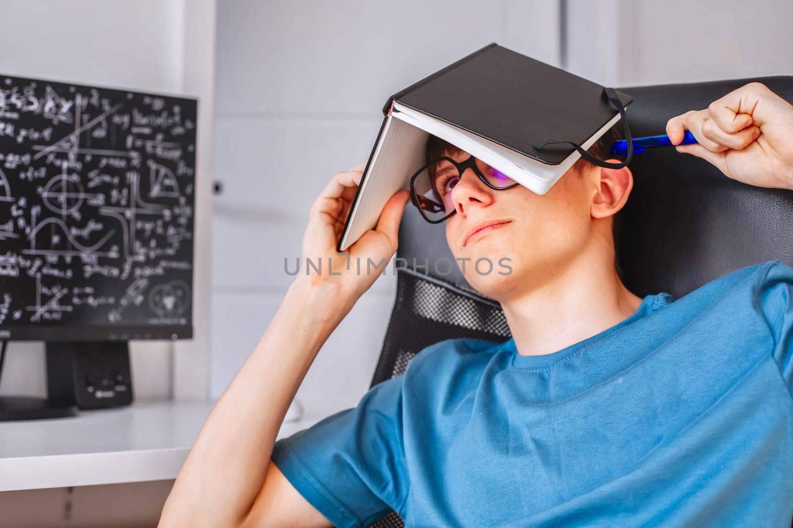 A teenage student wearing glasses sits at a computer with his head covered with a notebook, solving problems, preparing for an exam, mathematical symbols are displayed on the monitor. High quality photo