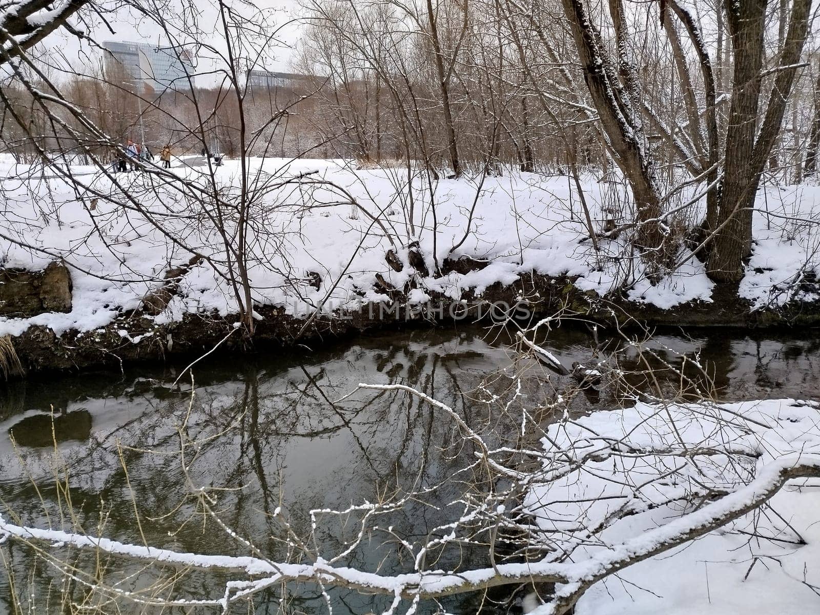 A river flows through a snow-covered park during winter. Bare trees line the riverbank, their branches reflected in the water. Distant figures walk through the snowy landscape in the background.