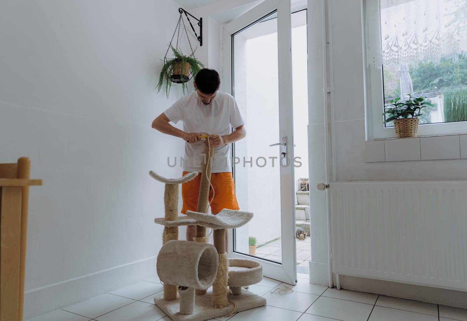 One young Caucasian handsome guy removes a jute thread, cutting with a construction knife from a cardboard tube of a cat's house to repair it, standing in the kitchen on a summer day, side view close-up with depth of field. Concept of repairing a cat's house at home.