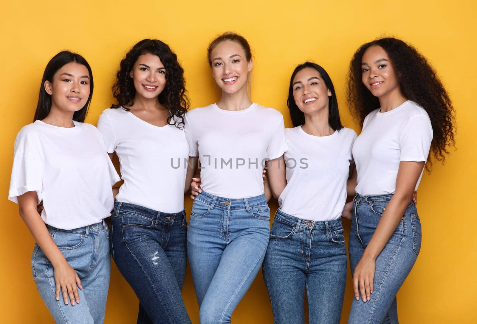 Group Of Five Diverse Models Ladies Posing Smiling To Camera Standing On Yellow Studio Background. Mixed Women Expressing Positive Emotions. Female Beauty And Diversity Concept
