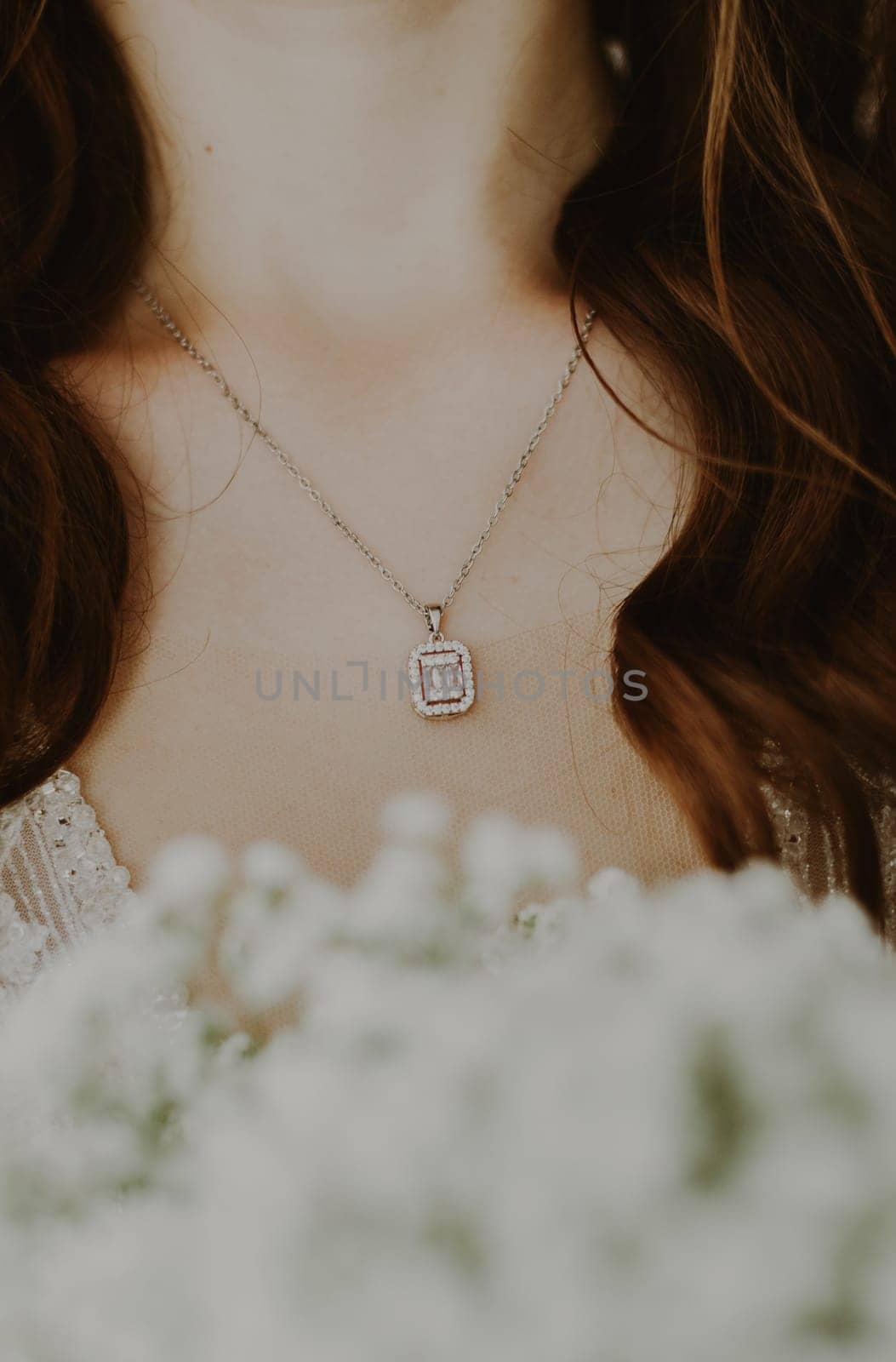 A beautiful chain with a precious pendant hangs on the neck of a young unrecognizable Caucasian brunette bride with a bouquet of white blurry boutonnieres standing on a summer morning in the room before the wedding ceremony, close-up side view.