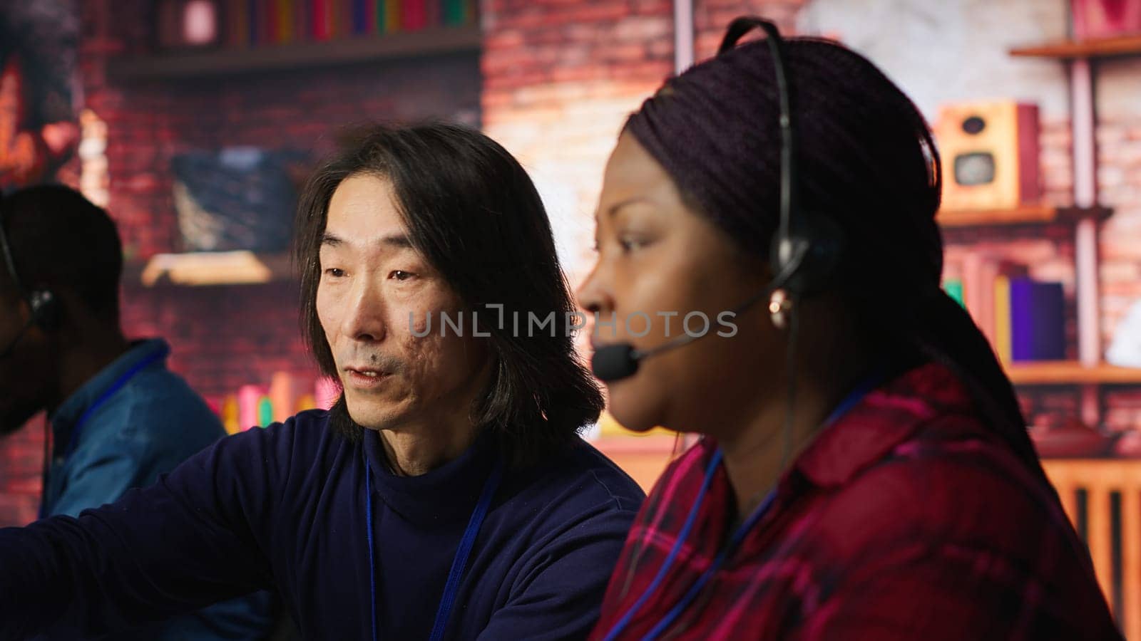 Experienced call center team leader assisting employee and drinking coffee in office. Asian account manager doing teamwork with colleague, enjoying beverage in workplace, camera A