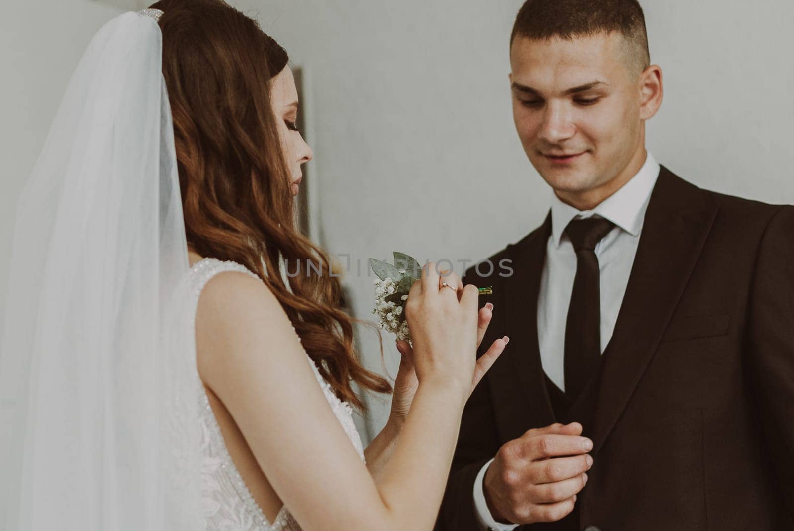 Portrait of one young beautiful stylish white brunette bride inserting a mini bouquet of boutonnieres into the chest pocket of the groom in a dark suit on a summer morning in the room, side view close up. Wedding traditions concept.