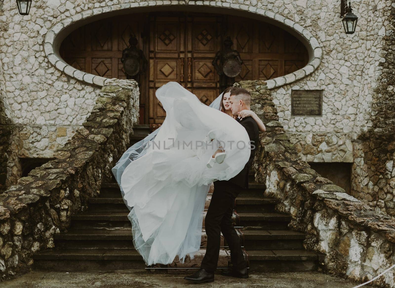 A young handsome Caucasian groom takes his bride in his arms to carry her to the car from the wedding castle on a cloudy rainy day, close-up side view with selective focus.