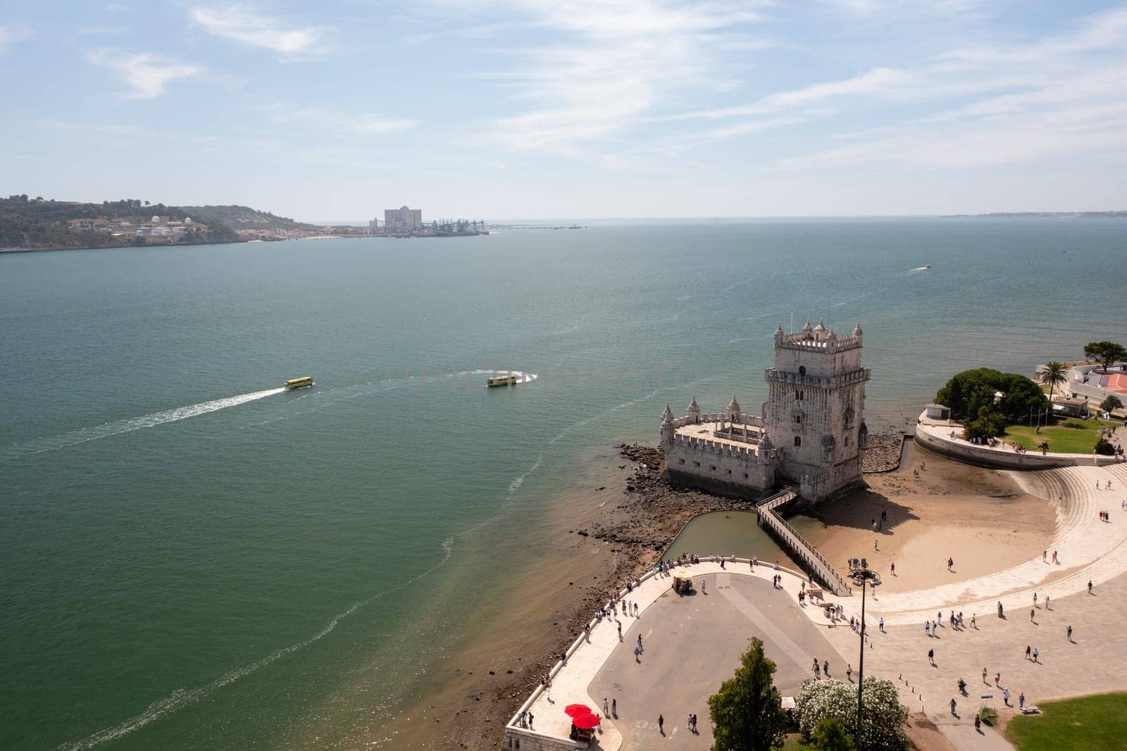 Aerial drone view of Belem Tower on Tagus River with boats creating wakes in the water, highlighting Lisbon's scenic waterfront and historic architecture