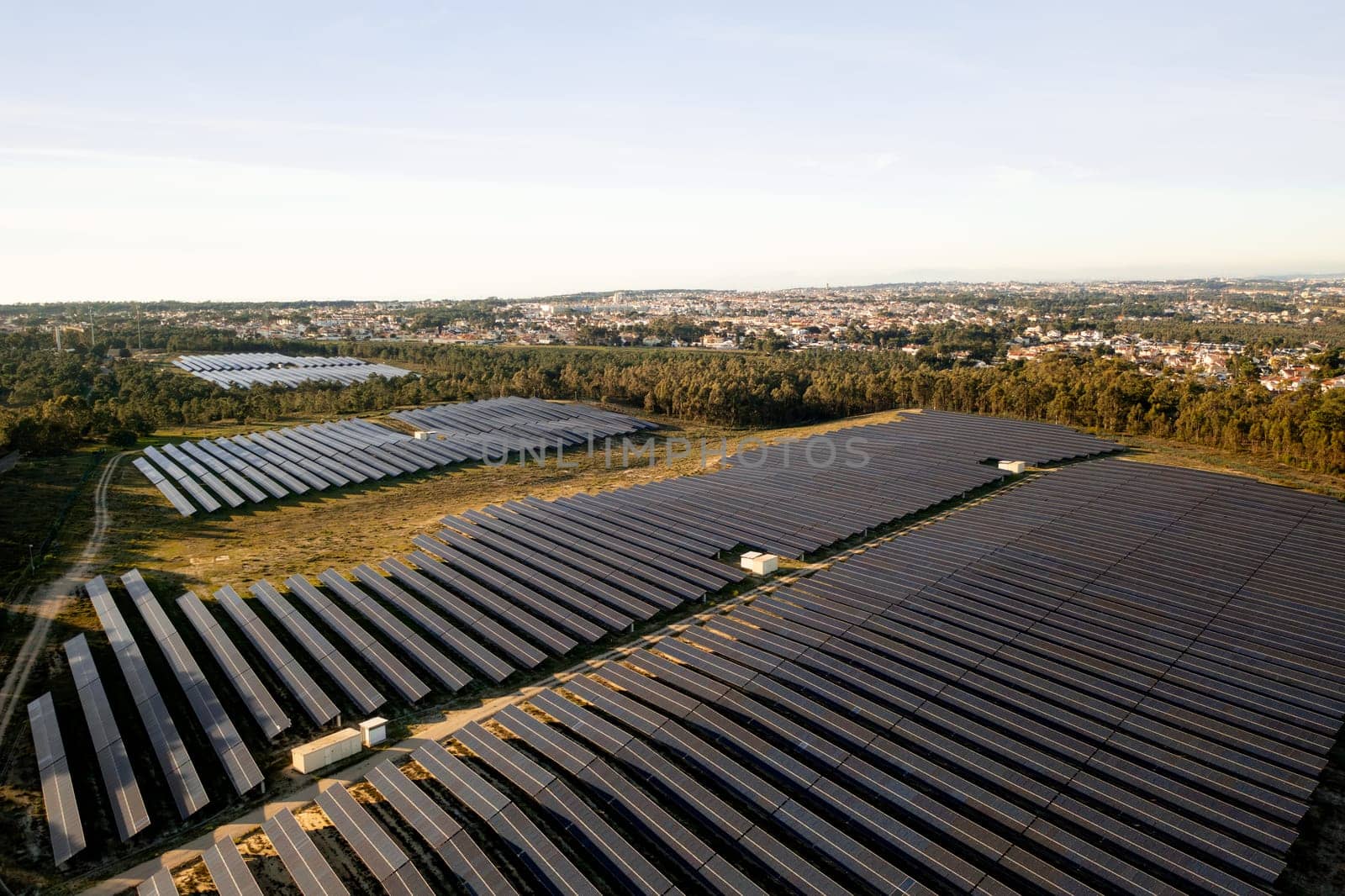 Solar farm with rows of solar panels covers large area surrounded by trees. Panels are arranged in parallel lines maximizing sunlight capture. Residential area is visible in background