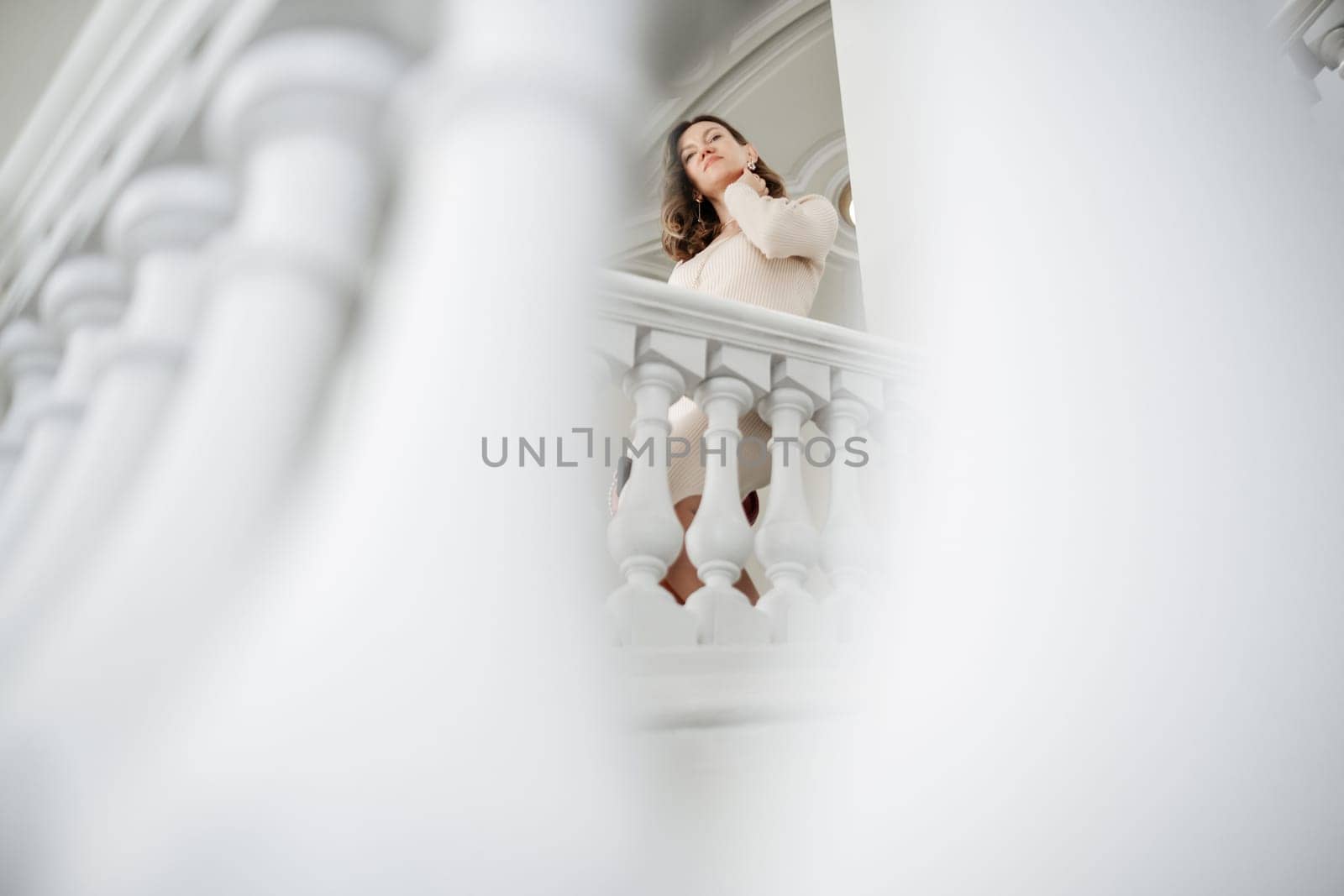A woman with long hair is standing elegantly on a white balcony, dressed in a stylish outfit, enjoying the bright daylight in a refined indoor space.