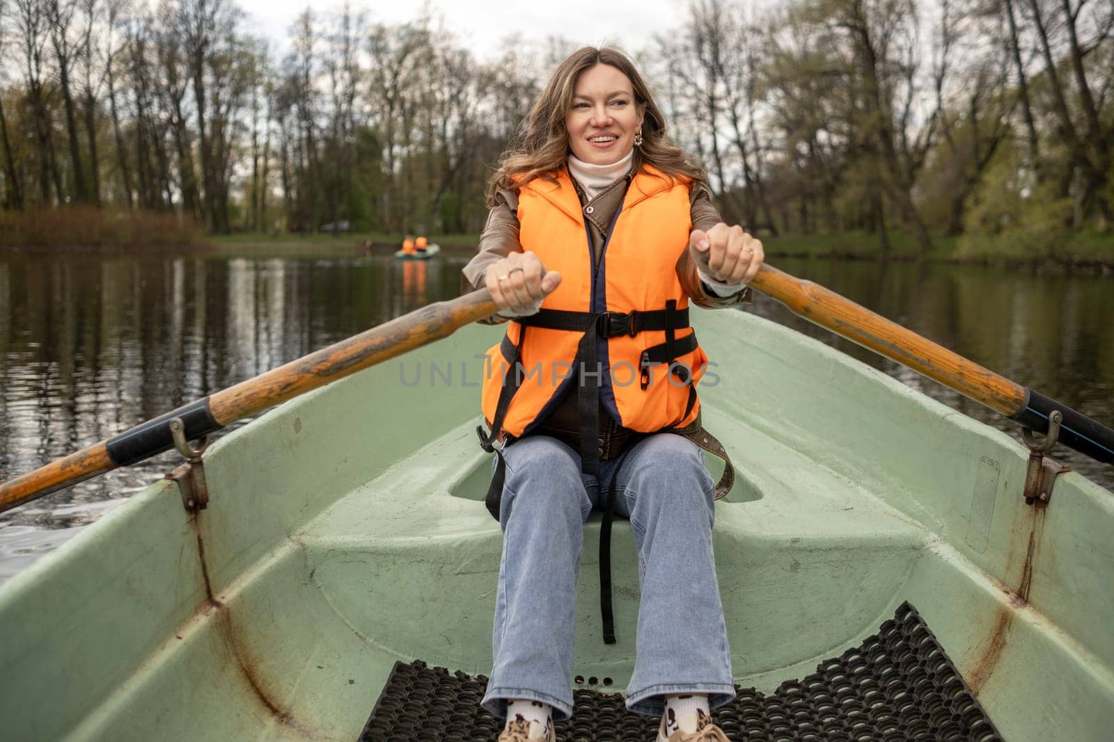 A smiling individual in a bright orange life jacket rows a small boat on a serene lake, with trees reflecting on the waters surface.