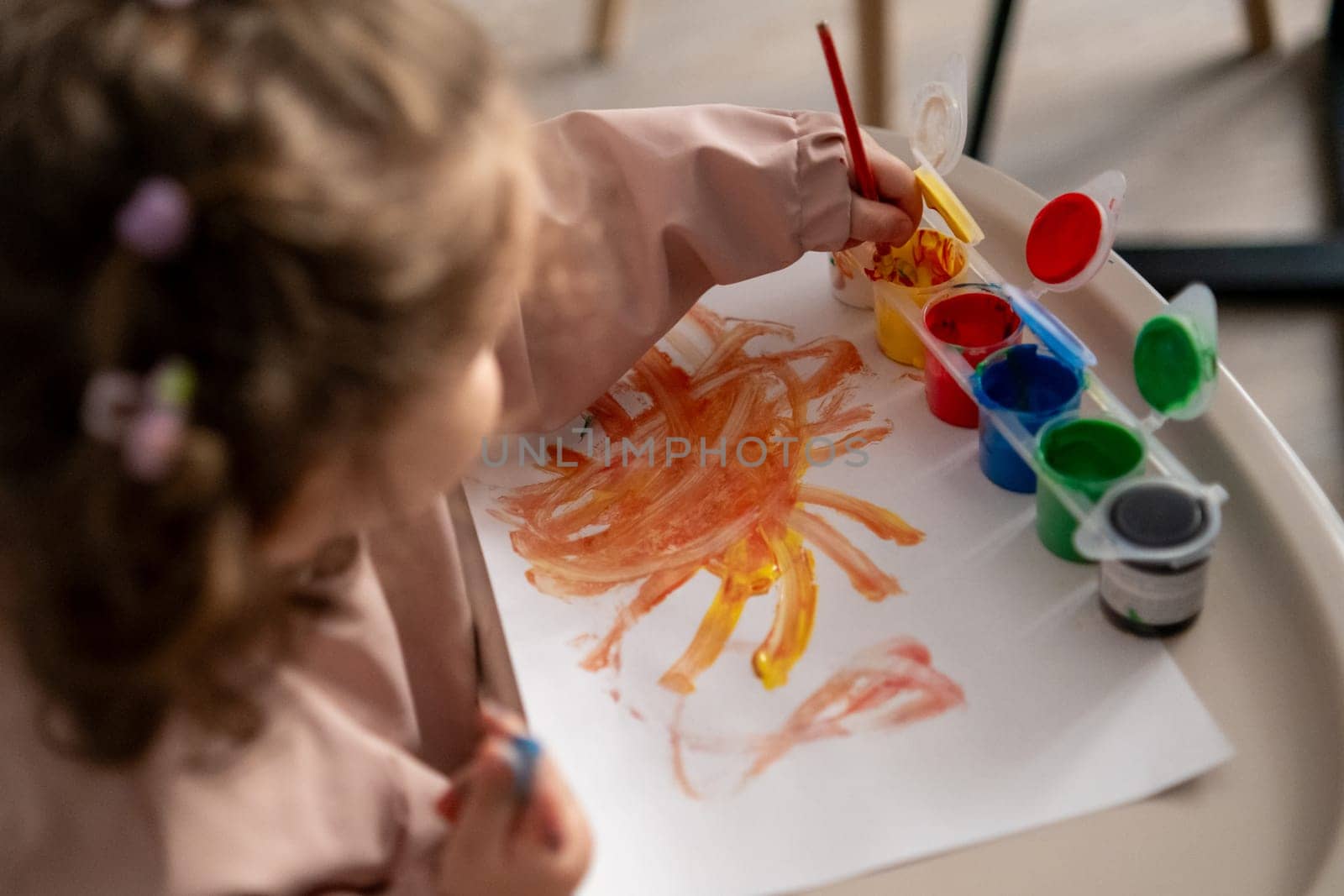 Focused little girl sitting at a kids table, carefully painting with a brush and gouache paints. Indoor creative activity, developing fine motor skills.