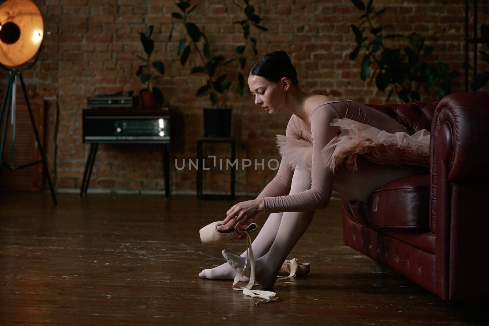 A poised ballerina is gently and delicately wearing her ballet shoes inside a beautifully stylish studio ambiance
