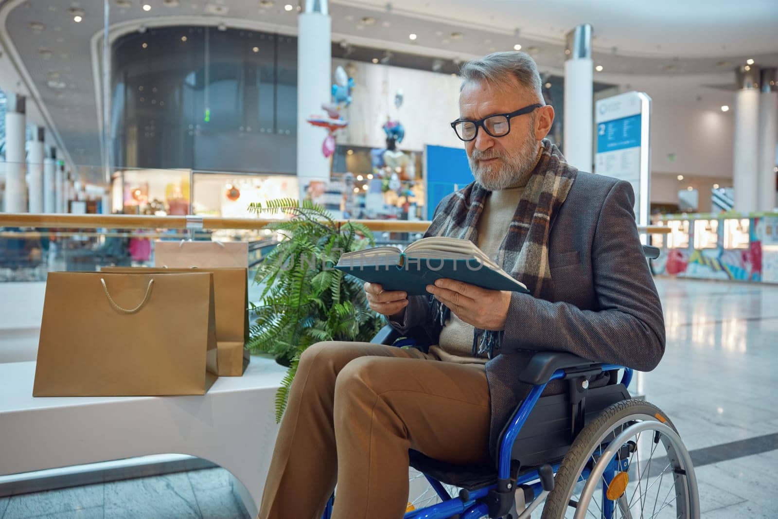 Man who uses wheelchair comfortably seated on bench and is engaged in reading an interesting book as he enjoys his day