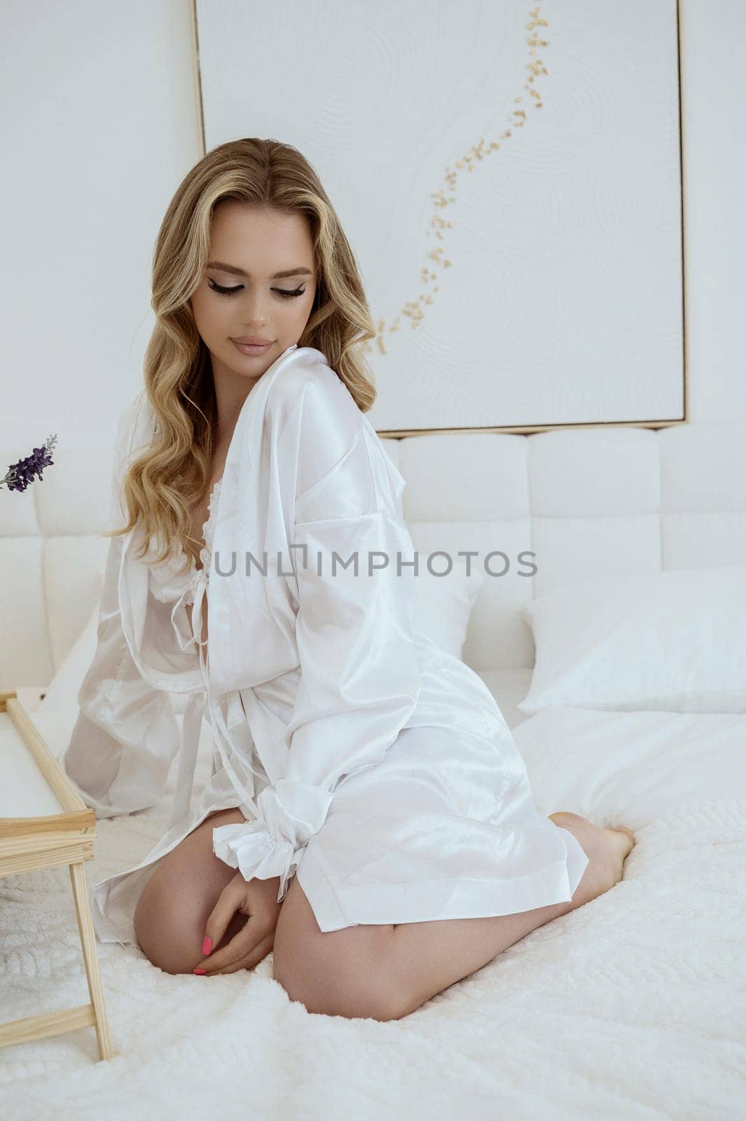 beautiful young blonde with long hair in silk white robe on bed. girl sits on white bed in light interior. she has clear skin. Domestic hair removal treatment concept