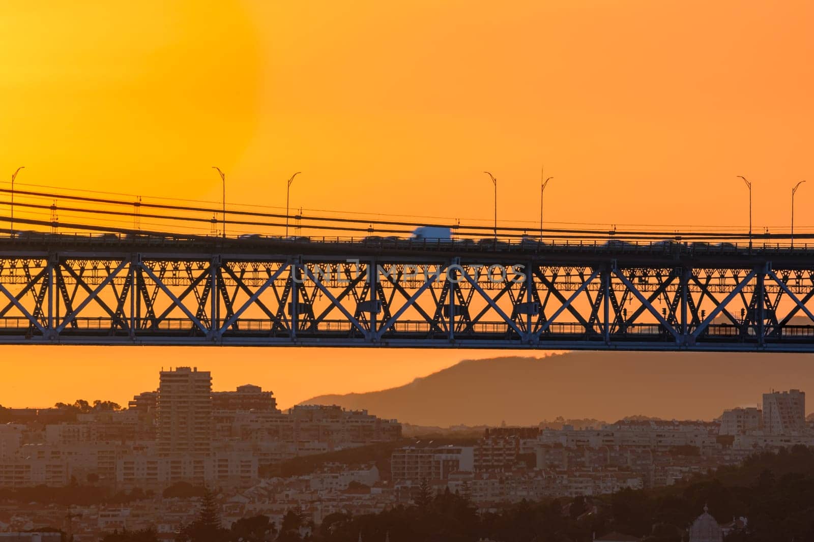 Beautiful view of golden sunset and suspension city bridge with its steel structure and cables, dramatic silhouettes of vehicles in motion blur against bright orange sky. Distant hills and urban skyline of modern buildings in evening summer haze.