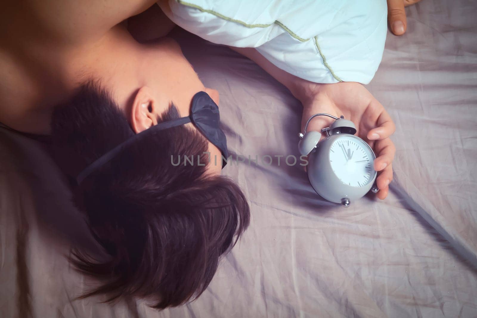 A young funny man sleeps soundly early in the morning, hugs a pillow and holds a vintage alarm clock in his hands, the sleep mask has slipped from his eyes.