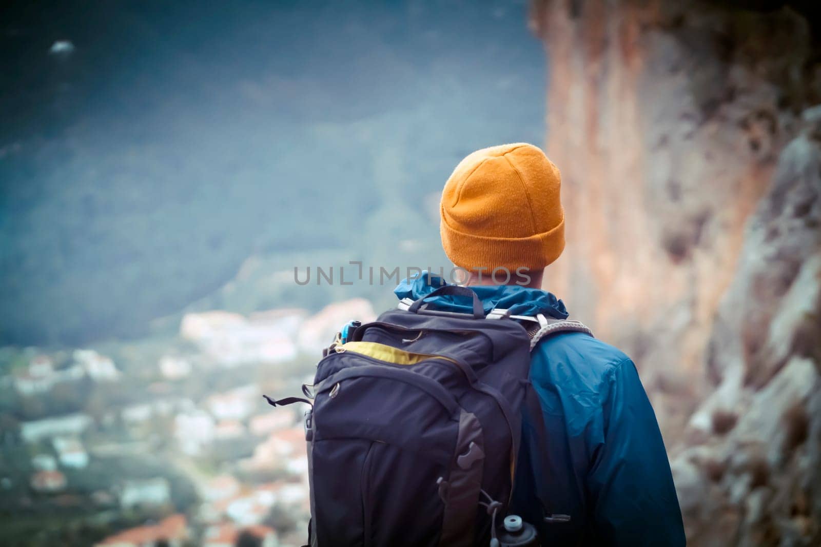 Young man in a yellow hat and with a backpack stands against the background of rocks and a view of a small village with red roofs, is engaged in trekking, discovers the beauty of the Mediterranean.