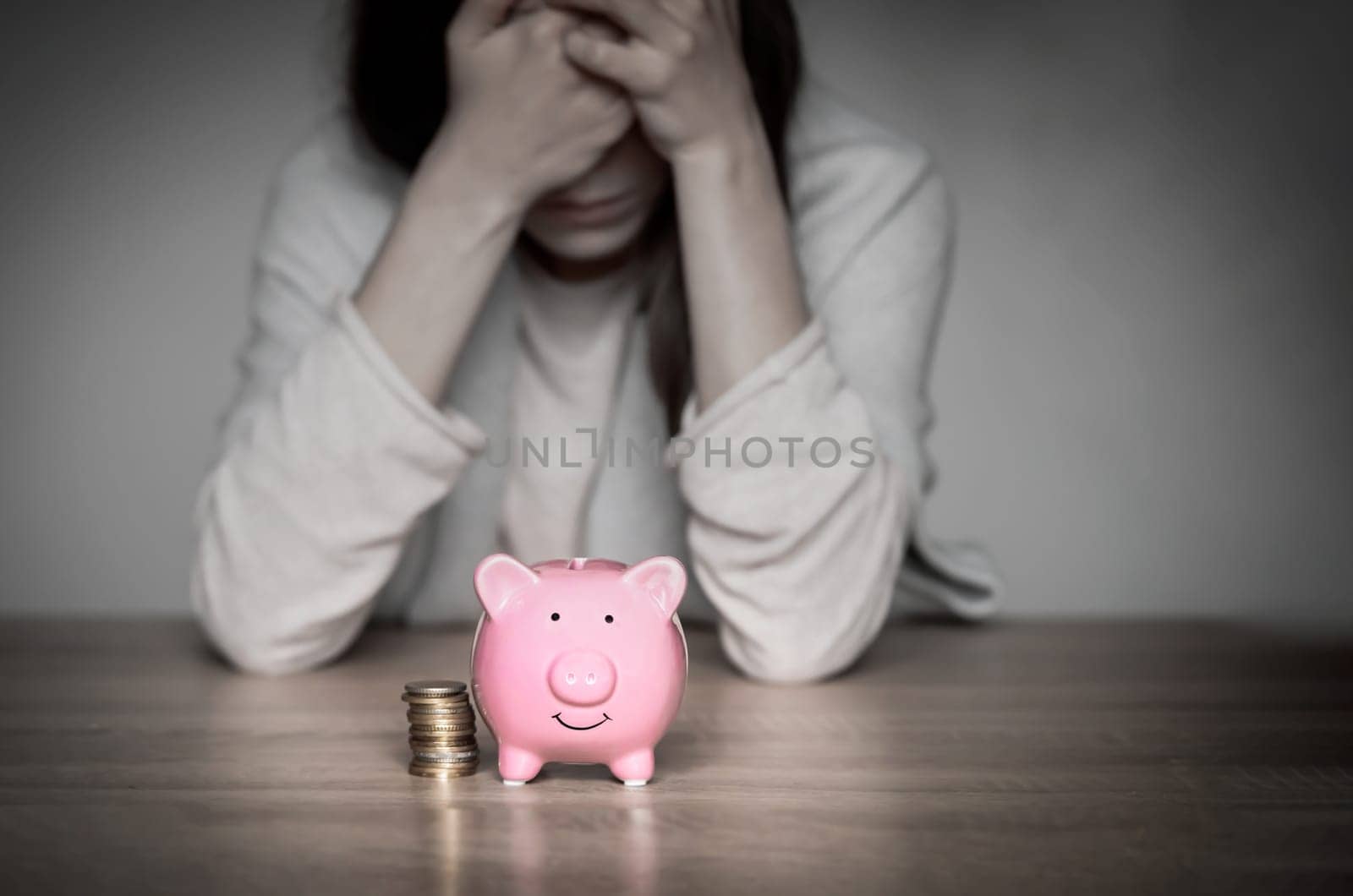 Sad girl leaned on the table, covering her face with hands, in the foreground a cute pink piggy bank. Woman lost her savings, was fired, and is experiencing financial difficulties, feels depressed.