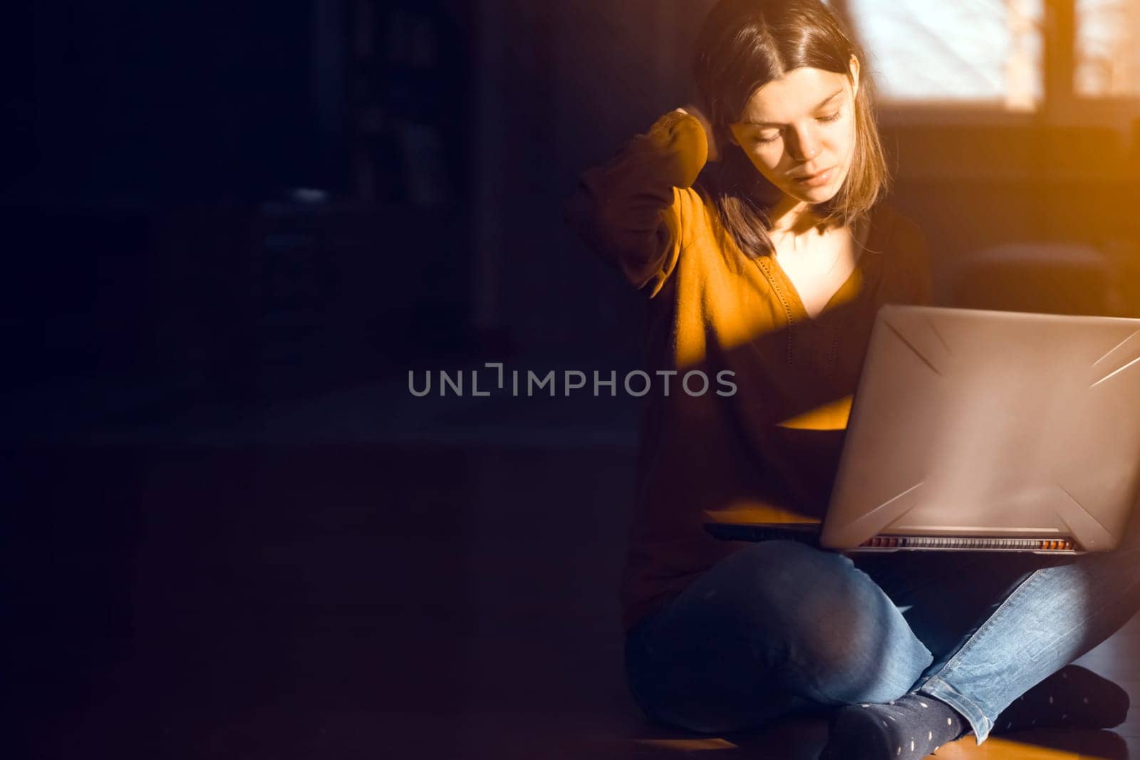 The girl sits on the floor of a cozy house in the rays of the sun and chats, works on a laptop online. A woman learns and develops her business at home.
