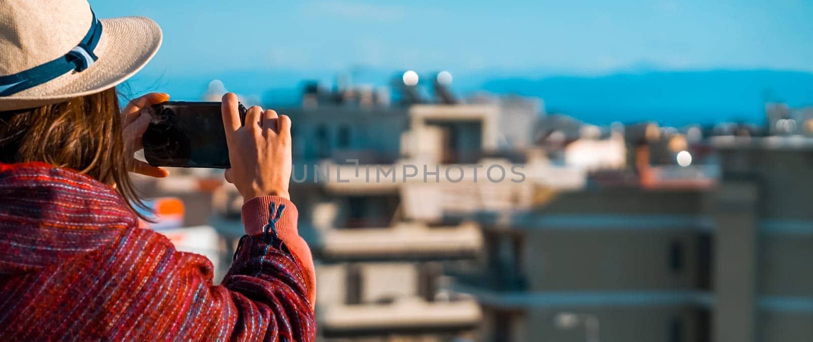 A young girl in a burgundy poncho and a hat travels and takes a photo with a cityscape on a sunny day.