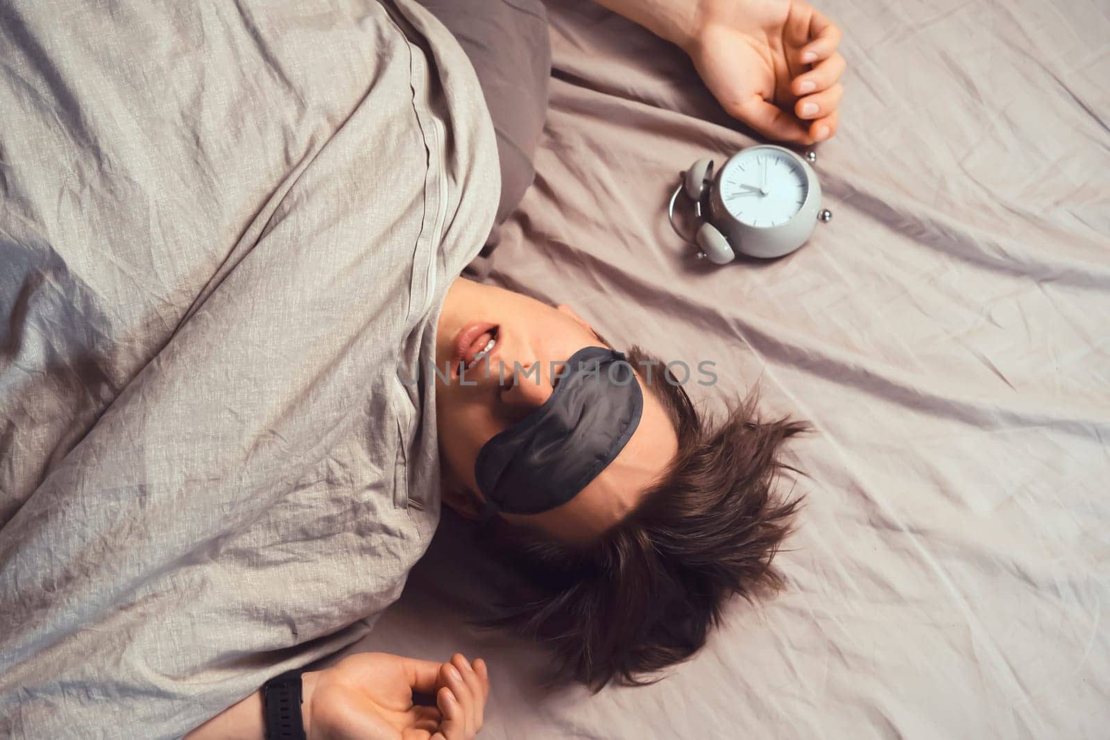 A young funny man sleeps soundly early in the morning, hugs a pillow and holds a vintage alarm clock in his hands, the sleep mask has slipped from his eyes.