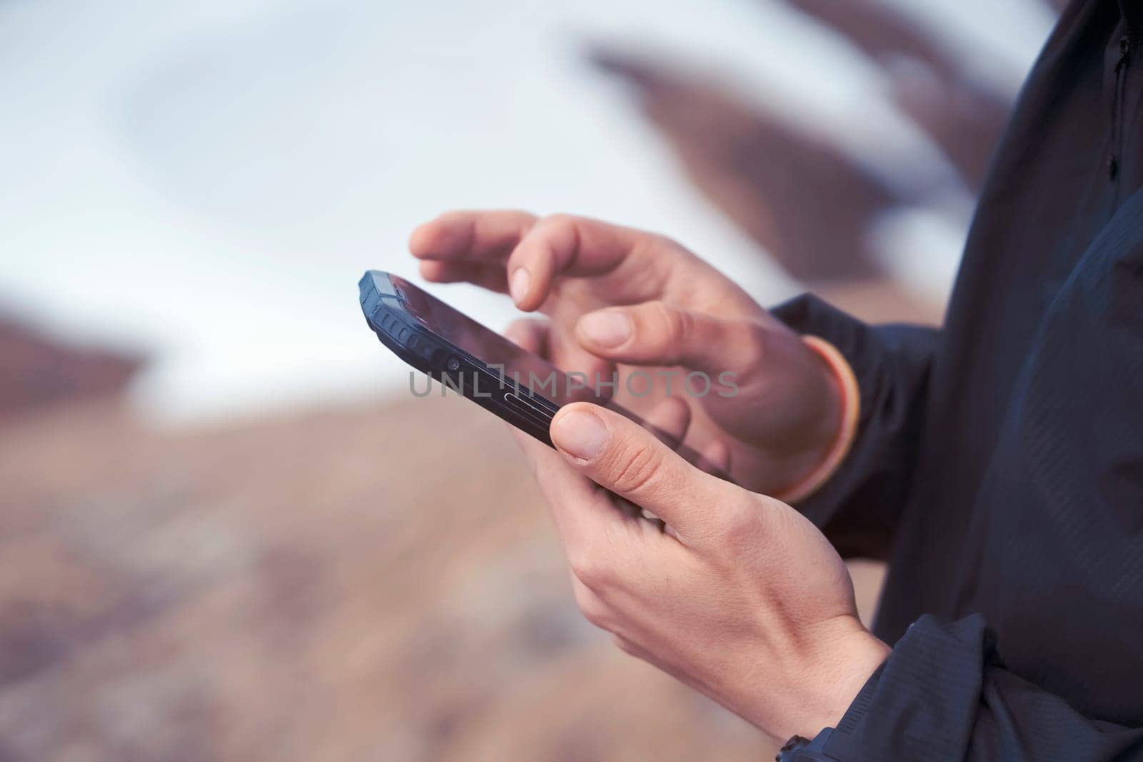 Man holds a smartphone in his hands and looks at a map, makes a route through the mountains, writes a message, chats. Young traveler explores the area, uses the app on his device while hiking.