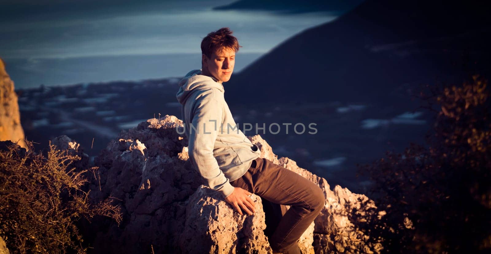 A man sits on a stone on top of a mountain in the sun, against the backdrop of a beautiful landscape with a sea coast and a small cozy city, the traveler is trekking during his trip.