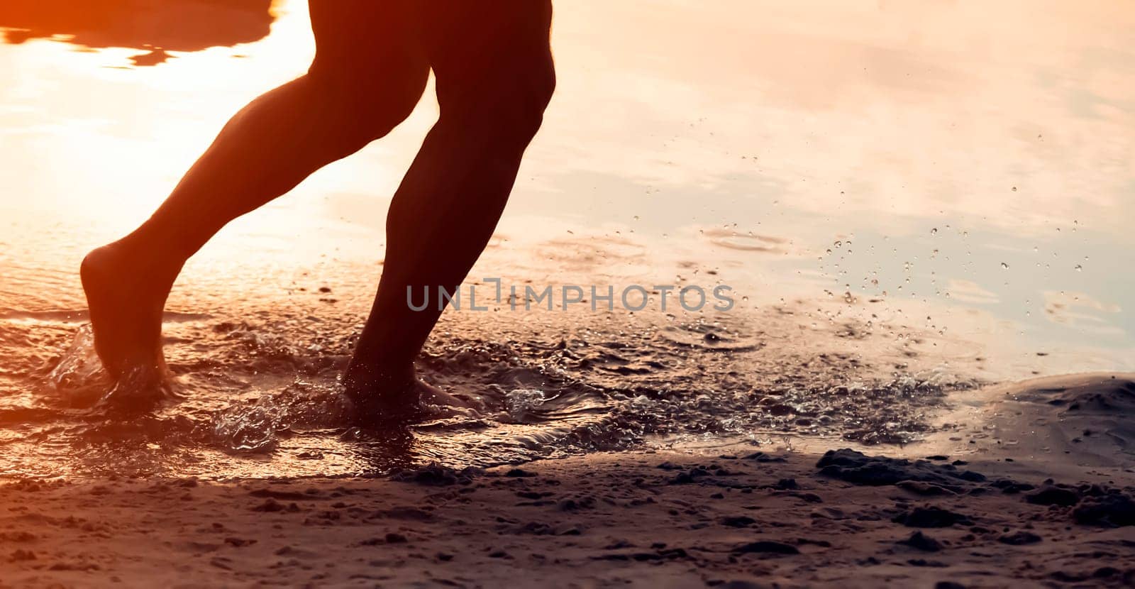 A person runs along the beach on the water, the sea or ocean in the rays of the sun, an athlete man spends time happily, is training, jogging outdoors, close-up view of the legs and splashes.