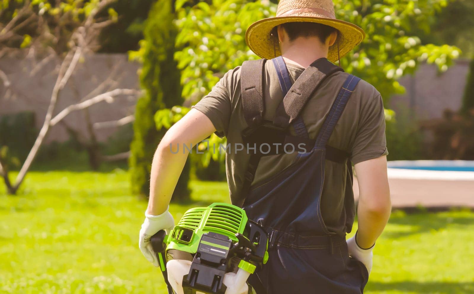 A young man in a straw hat is mowing a lawn with a lawn mower in his beautiful green floral summer garden. A professional gardener with a lawnmower cares for the grass in the backyard.