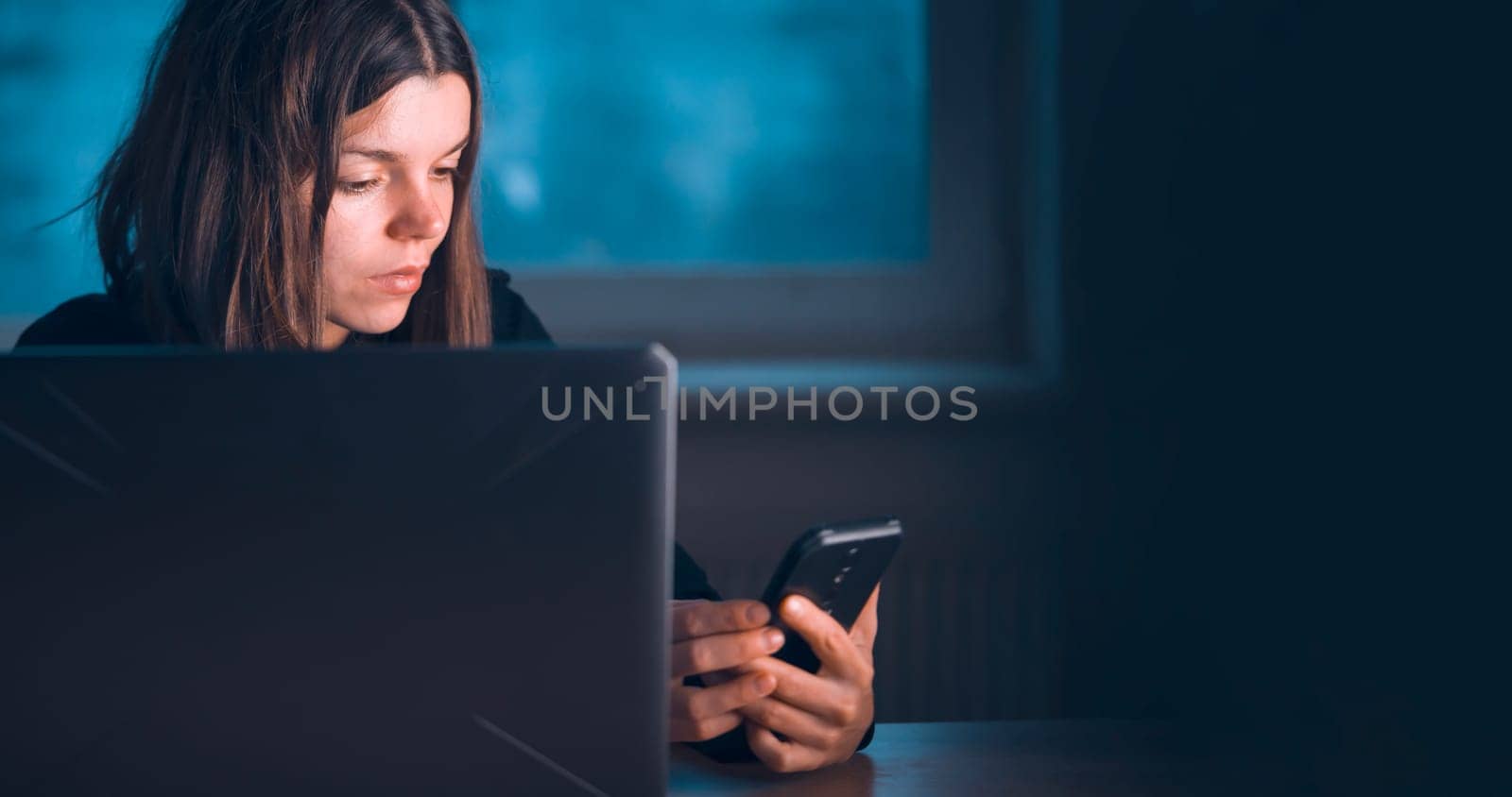 A young girl works at home at night with a laptop, a woman holds a phone in her hands, participates in a conference meeting online, studies, searches for information using her device.