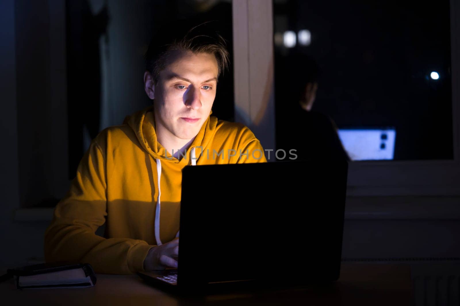 A young man sits and works at night on a laptop on the network, types on the keyboard, writes text messages, studies, prepares a project, a programmer, web designer works as a freelancer at home.