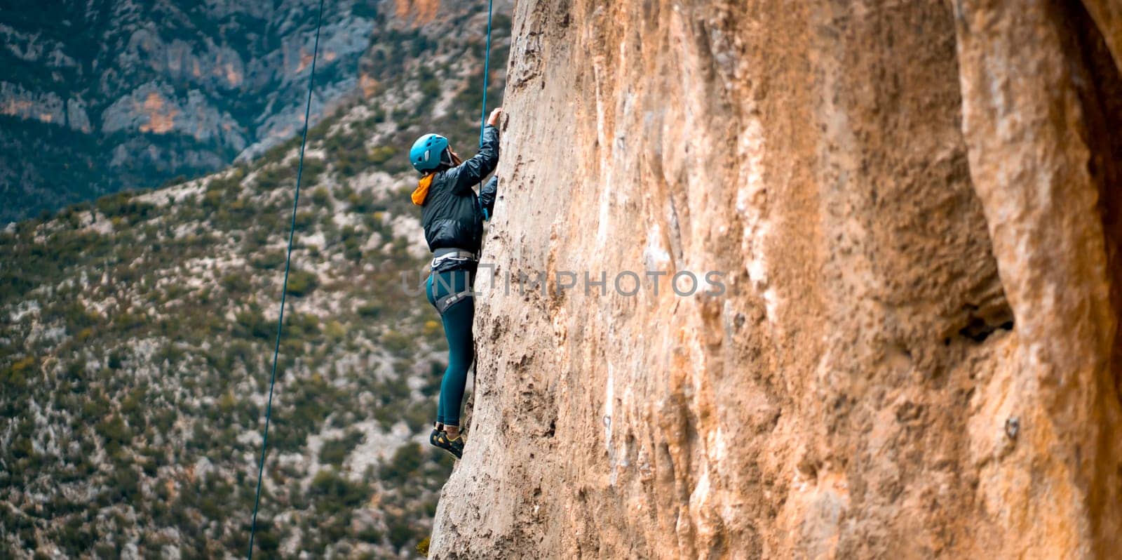 Young girl is engaged in extreme sports, mountaineering, leads an active lifestyle, climbs a rock with climbing equipment, a rope and in a helmet, against the background of hills with olive trees.