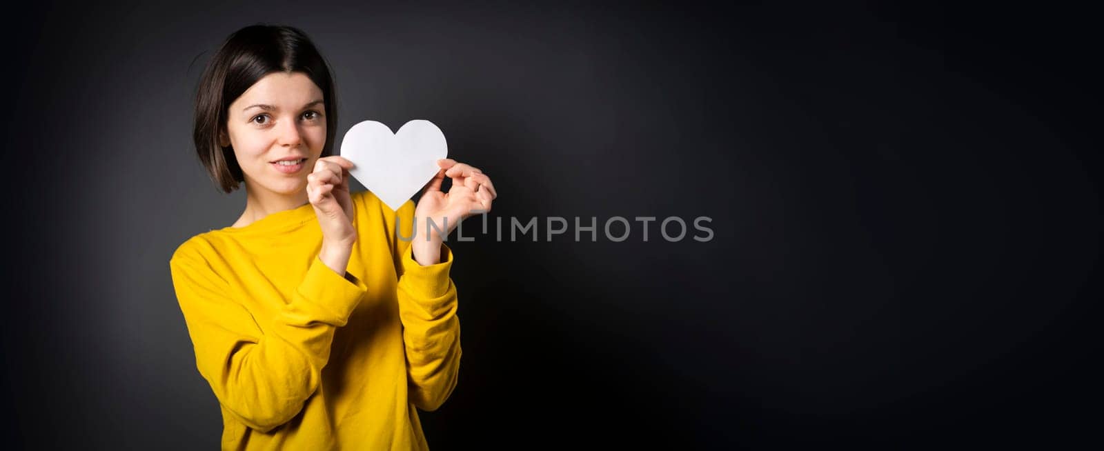 A young girl holds a white paper heart - a postcard or shows feelings of love. A woman with a joyful surprised face before the holiday and in search of love, relationships or shares her impressions.