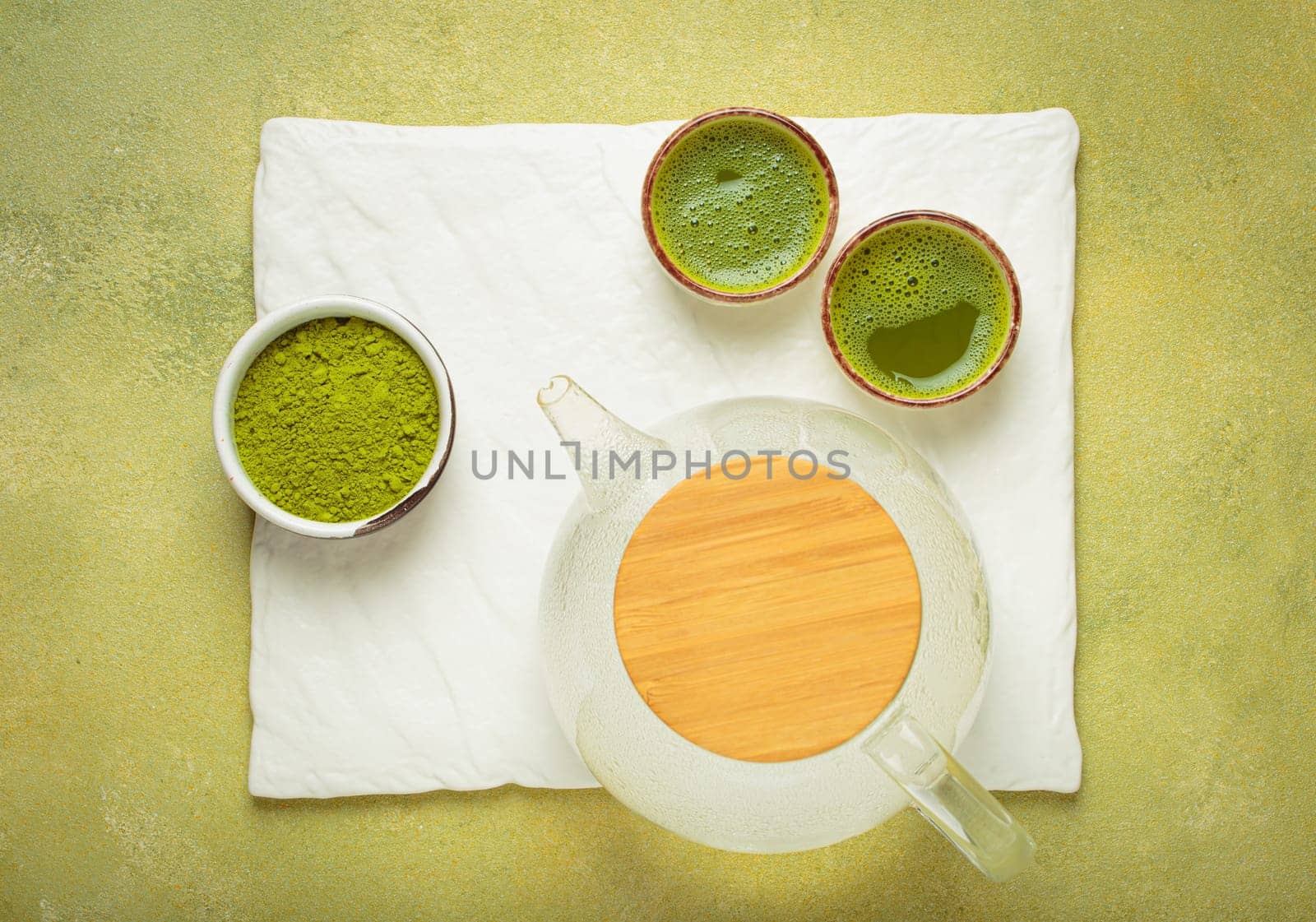 Glass teapot with wooden lid, two cups of matcha tea and bowl of matcha powder are laying on white stone slate on a green background, creating a relaxing and refreshing atmosphere