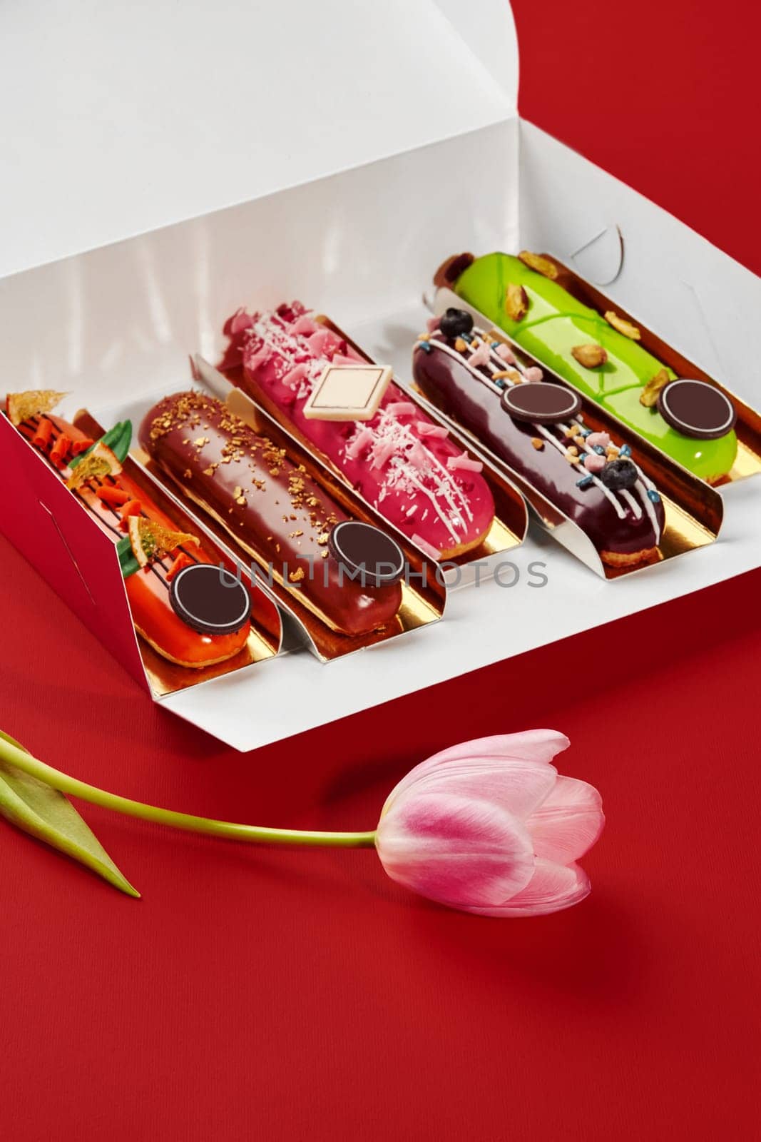 Set of colorful eclairs in open box with pink tulip on red background, celebrating a special occasion