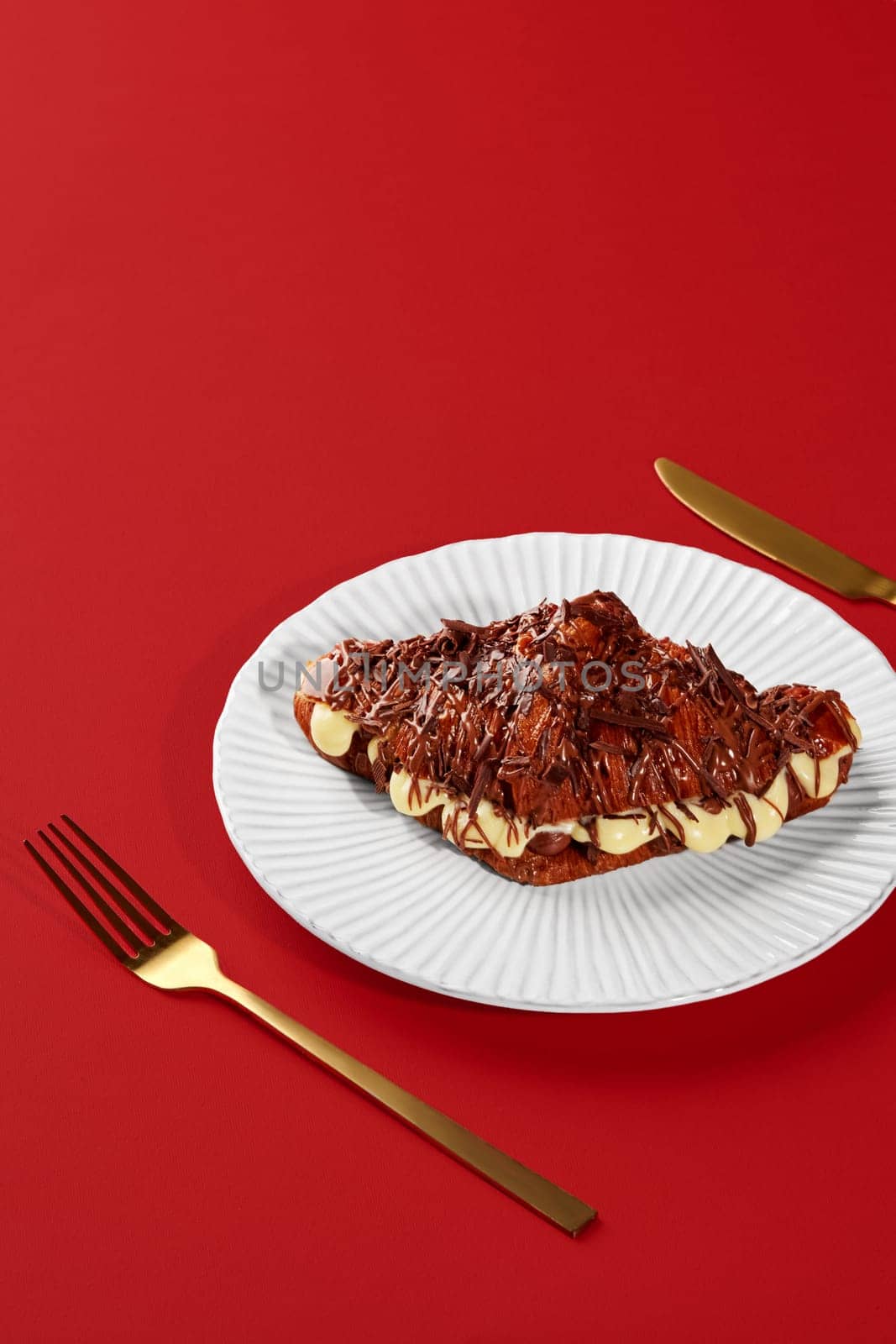 Chocolate covered croissant filled with cream served on white plate with golden fork and knife on vibrant red background