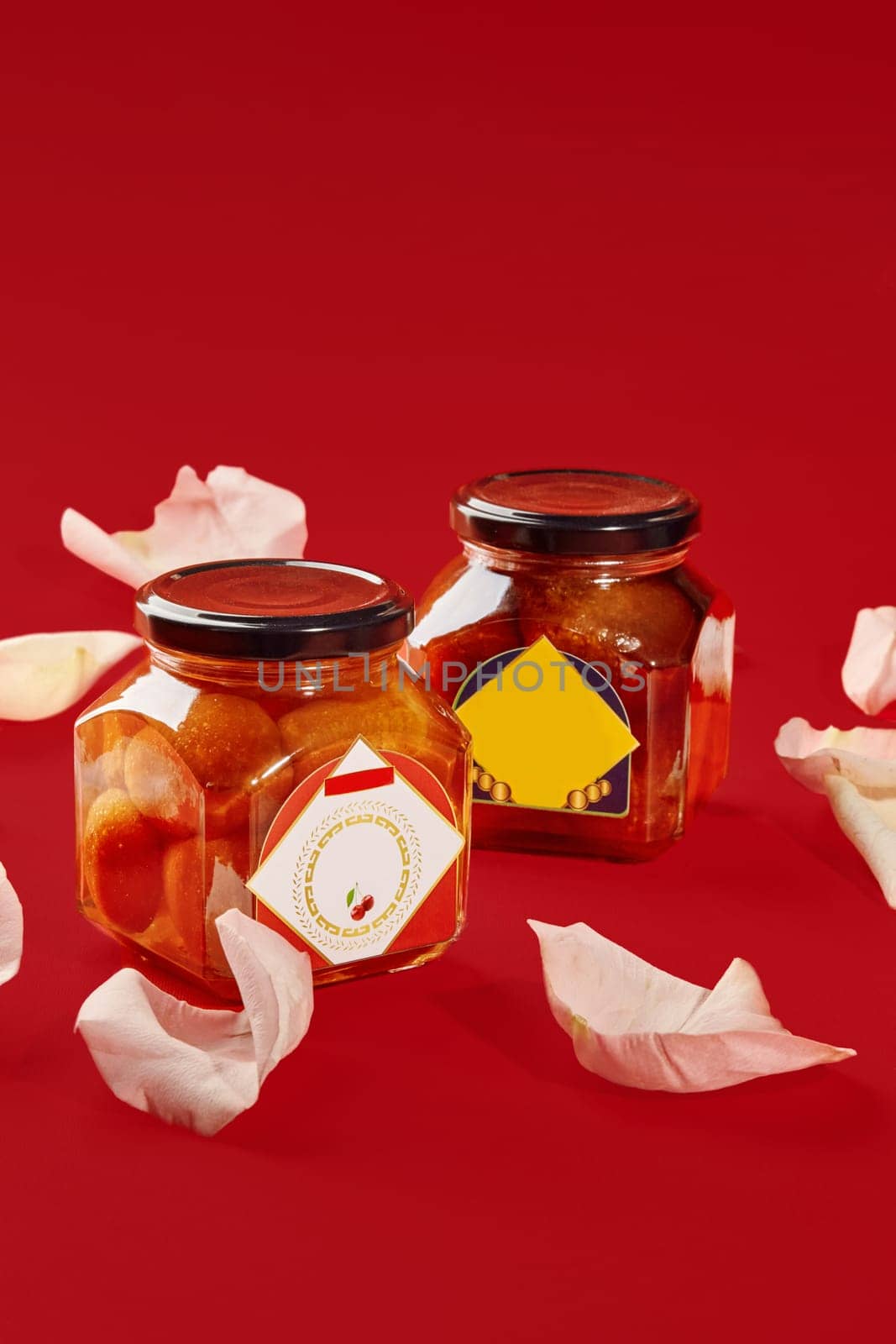 Two luxardo baba al maraschino jars surrounded by rose petals on a vibrant red background, creating a luxurious and romantic atmosphere