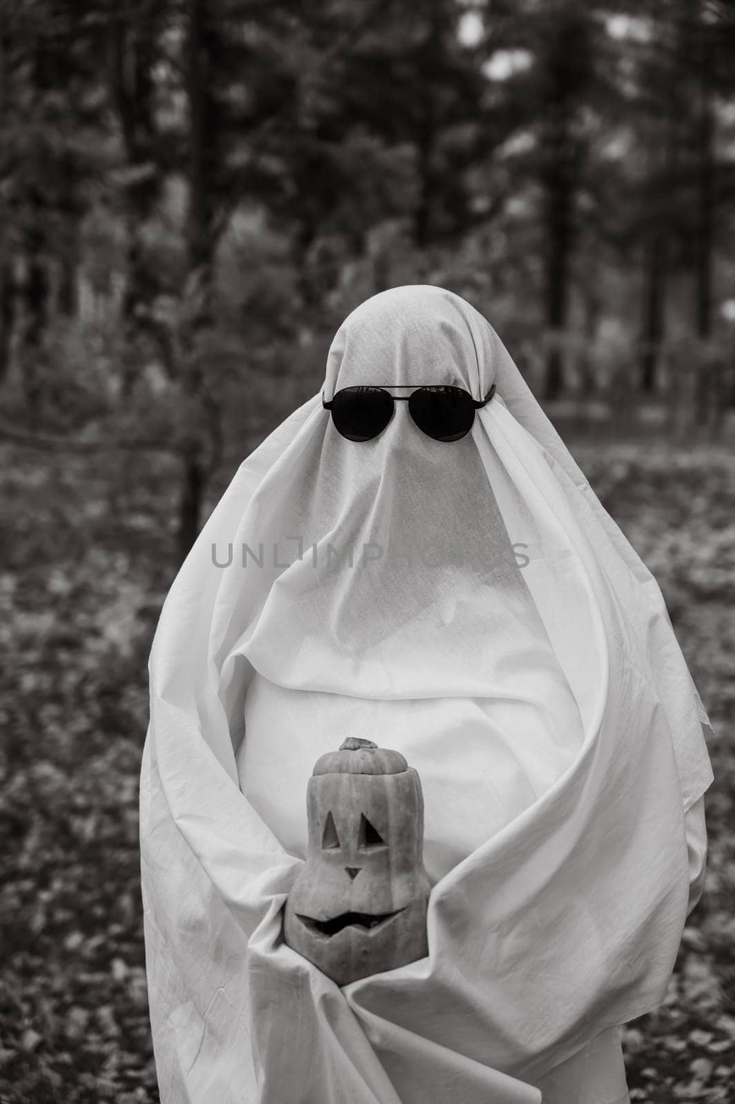 Woman in white sheet holding jack o'lantern against autumn forest background. Halloween costume. Black and white photo