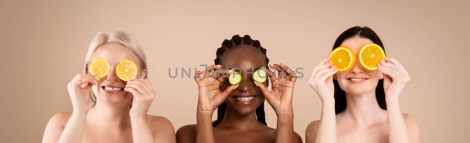 Beauty And Skin Care. Three Diverse Women In Bath Towels Covering Eyes With Cucumber And Orange Slices, Making Organic Face Masks, Posing Together Over Beige Studio Background, Copy Space