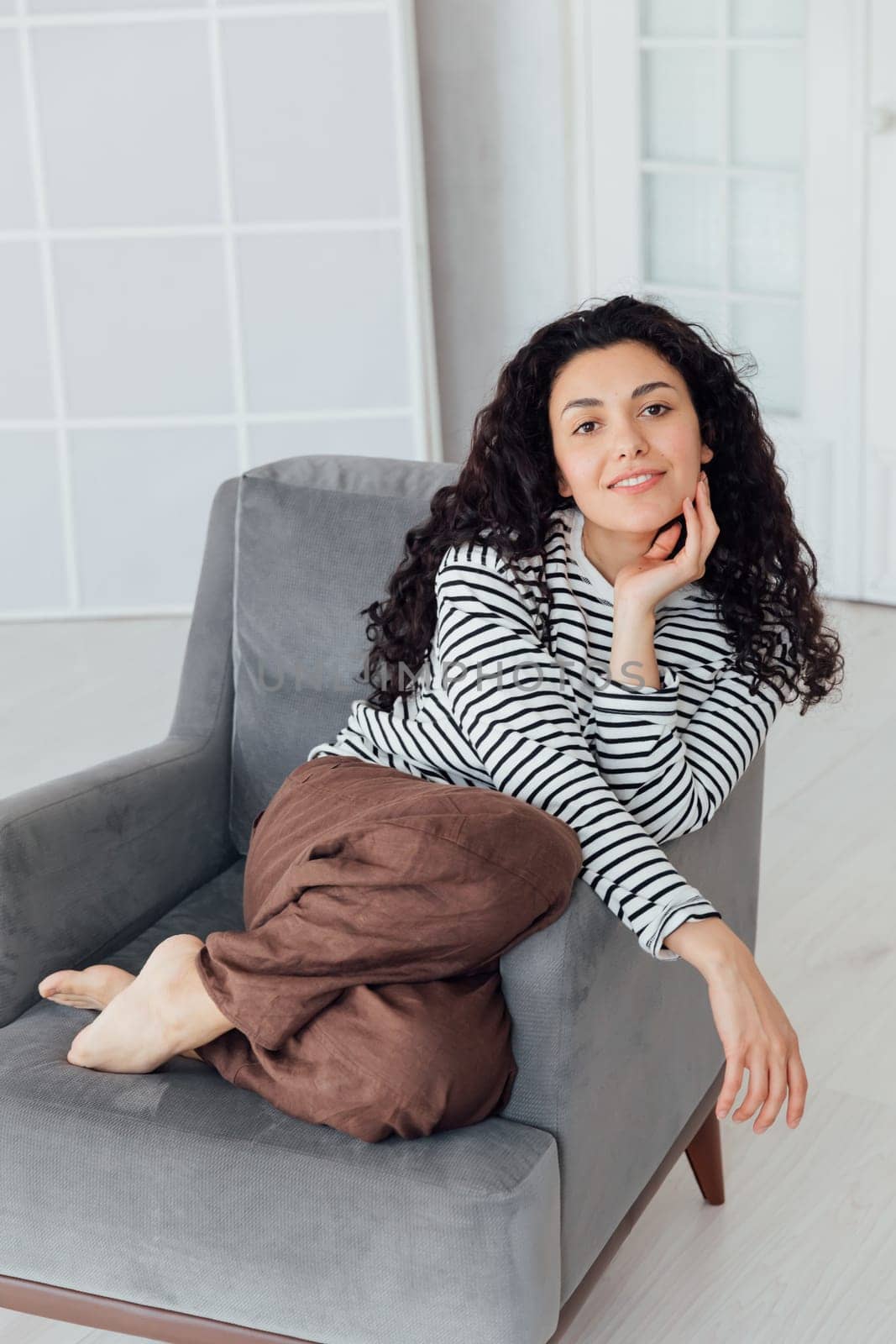 Curly brunette woman in homemade coat sitting on armchair smiling