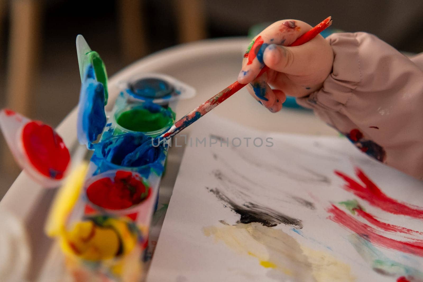 Close up of a toddlers hand reaching for a paintbrush with gouache colors. Creative activity at home, early childhood art education.