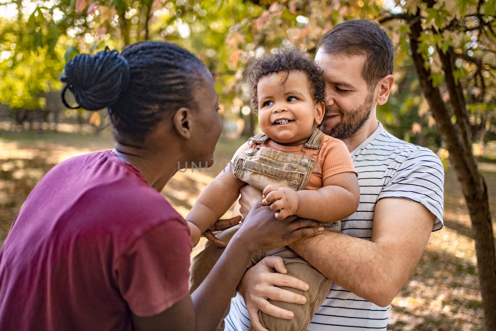 Two parents embrace their joyful baby boy in a vibrant park. Sunlight filters through colorful trees, creating a warm, loving atmosphere as laughter fills the air.