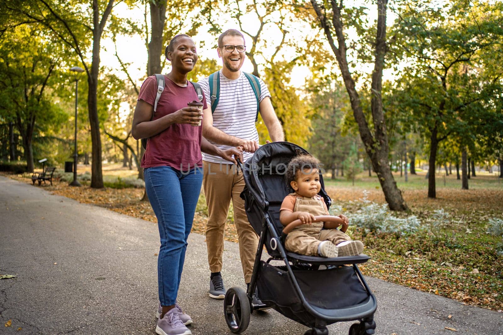 A couple enjoys a leisurely walk in a vibrant park, surrounded by autumn leaves. Their playful baby in a stroller observes the beautiful day, creating a warm family moment.