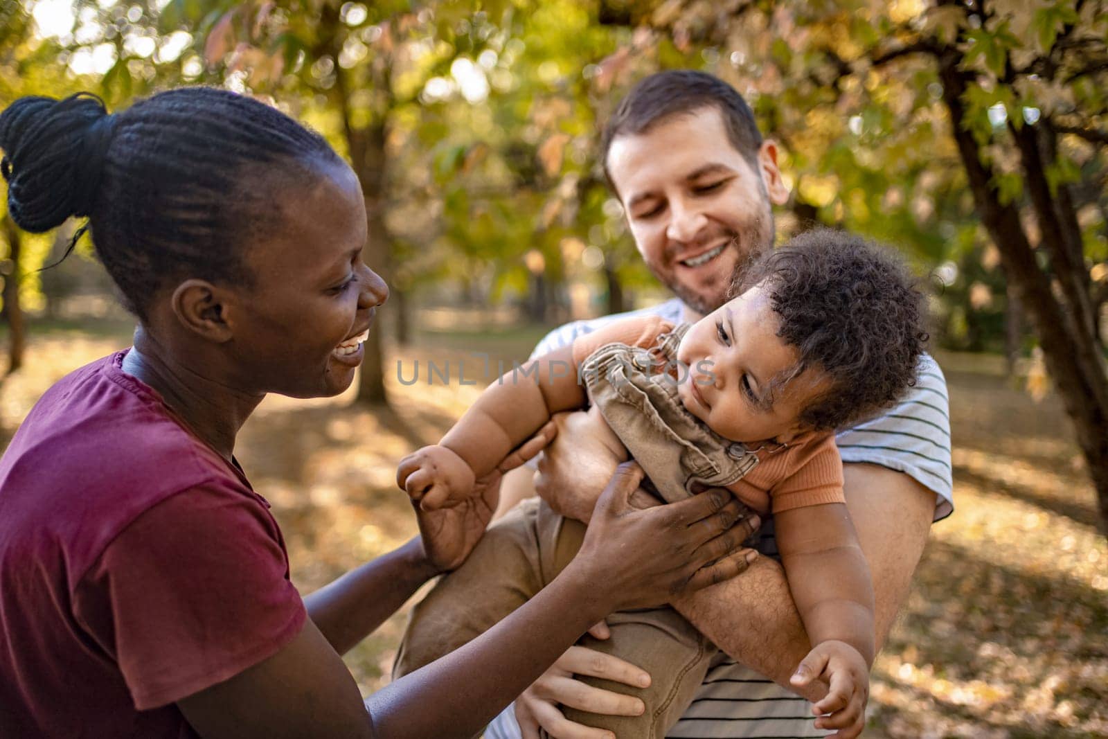 In a serene park filled with autumn foliage, a joyful couple enjoys playful moments with their curious baby. Warm sunlight filters through the trees, creating a cozy atmosphere.